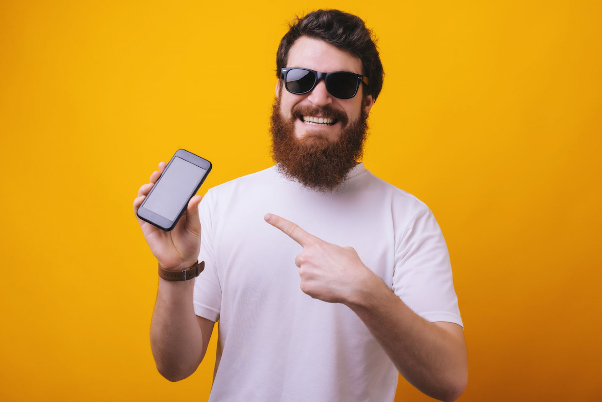 Image of young brunette bearded man in sunglasses pointing finger on smartphone and smiling on camera